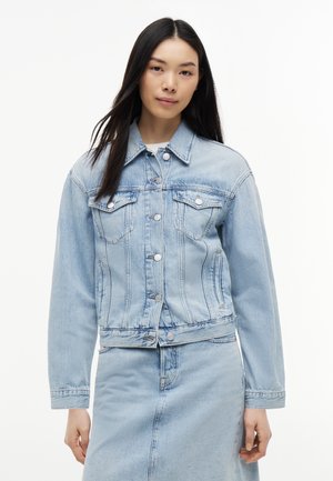 Junge Frau mit langem dunklem Haar, die eine hellblaue Jeansjacke und einen passenden Rock trägt, steht vor weißem Hintergrund.