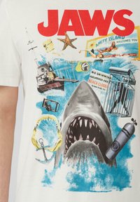 T-shirt en coton blanc avec le texte "JAWS" en rouge, une illustration de requin, un équipement de plongée, des bouées de sauvetage et des vagues océaniques en couleurs bleue et verte.