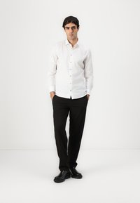 White long-sleeve shirt with button front, paired with black trousers and black shoes. Simple design, no visible patterns or textures.