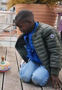 Un colorato yo-yo in legno su un pavimento in legno con un bambino inginocchiato, che indossa un giubbotto verde e un maglione blu, concentrato sul giocattolo.