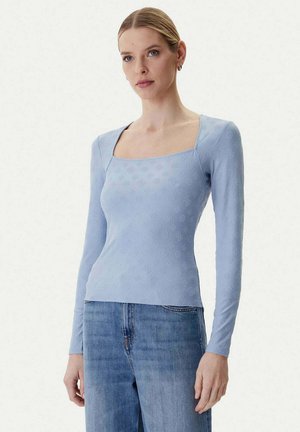 Donna che indossa una maglia aderente a maniche lunghe di colore azzurro chiaro con un motivo sottile e scollo quadrato, abbinata a jeans blu a vita alta.