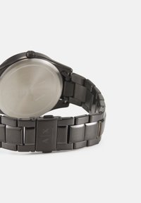Un orologio in metallo nero con un bracciale testurizzato, una cassa tondeggiante e un retro liscio e riflettente di colore argento con dettagli del logo incisi.