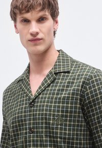 Camisa a cuadros verde y beige con cuello con muesca, que incluye un bolsillo, cierre de botones y un logo bordado en el pecho.