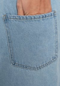 Lichtblauwe denimzak met een gladde textuur, voorzien van contrasterend stiksel. Een hand steekt gedeeltelijk in de zak.