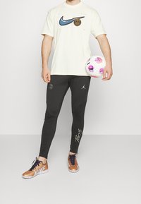 Biała koszulka z krótkim rękawem z niebieskim logo Nike, czarne zwężane spodnie z emblematem Paryża i pomarańczowe buty Nike z białymi akcentami, trzymająca wzorzystą piłkę nożną.