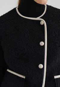 Veste noire à texture, dotée d'un col montant, de passepoils beiges et de grands boutons. Comprend deux poches latérales avec des accents contrastants.