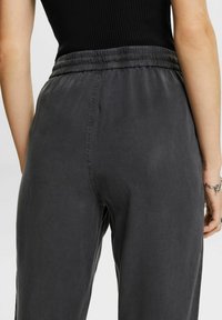 Gros plan sur une personne portant un pantalon gris foncé taille haute, ample, avec une taille élastique, et un haut sans manches noir.