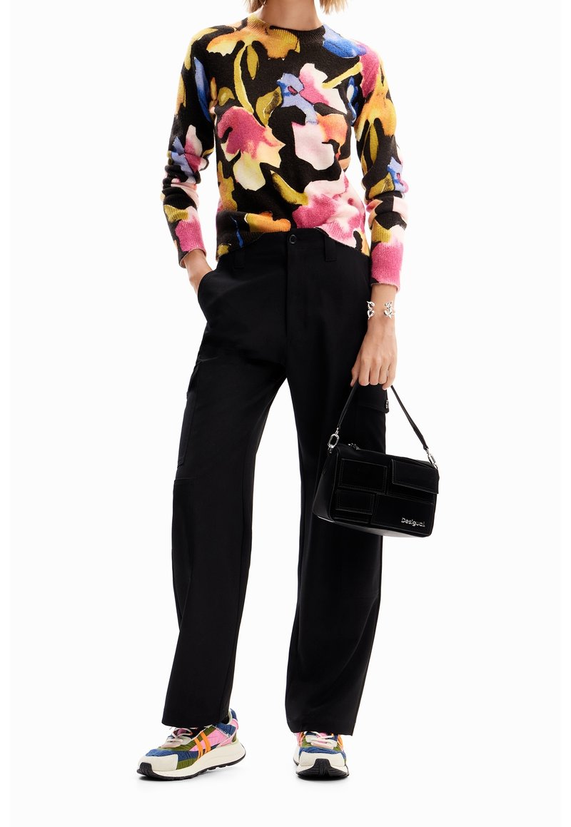 Personne portant un pull coloré à motifs floraux, un pantalon noir à jambes larges, des baskets multicolores, et tenant un petit sac à main noir.