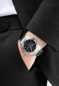 Reloj de acero inoxidable plateado con una esfera negra, acentos azules, tres subesferas de cronógrafo y una correa pulida, llevado en una mano con un traje negro.