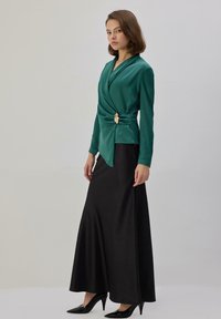 Mujer con blusa cruzada color verde azulado con un broche dorado y falda larga negra, usando tacones negros, posando frente a un fondo claro y liso.