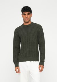 Maglione lavorato a maglia color verde oliva con un motivo testurizzato, dotato di collo rotondo e polsini e orlo a coste, abbinato a pantaloni bianchi.