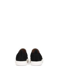 Zapatos negros de ante tipo slip-on con suela de goma blanca, que presentan un diseño minimalista y una textura suave. La vista trasera resalta los detalles de las costuras.