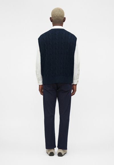 Άτομο με κοντά ξανθά μαλλιά που φοράει ένα μπλε σκούρο πλεκτό γιλέκο πάνω από ένα λευκό πουκάμισο, σκούρο παντελόνι και μπεζ παπούτσια, με την πλάτη γυρισμένη σε απλό φόντο.