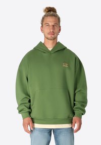 Grüner Kapuzenpullover mit lockerer Passform, ausgestattet mit einer Fronttasche und einem kleinen Emblem auf der Brust. Weicher Stoff und gerippte Bündchen.