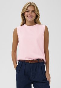 Bez piedurknēm rozā kokvilnas topiņš ar apaļu kakla izgriezumu, brīvs piegriezums un sānu apmale. Pāris ar augsto vidukli džinsu biksēm un brūnu jostu.