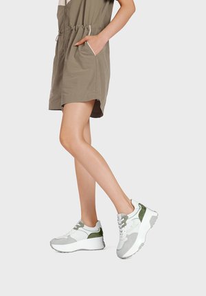 Persona che indossa un vestito kaki corto con coulisse in vita e sneaker bianche con dettagli verdi e grigi, in piedi contro uno sfondo semplice.