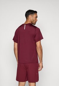 Bordový športový top s krátkymi rukávmi, okrúhlym výstrihom a reflexným akcentom na chrbte, v kombinácii s odpovedajúcimi šortkami. Hladká textúra, moderný strih.