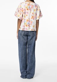 Donna che indossa una blusa a maniche corte floreale con colori pastello e jeans blu chiaro larghi, in piedi di spalle.