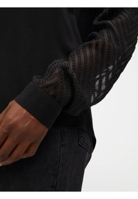 Top negro con mangas transparentes texturizadas con un patrón geométrico y un puño negro sólido. La tela parece suave con un ligero brillo.
