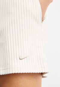 Vit ribbad tygkortärmad skjorta med rundad fåll. Har en liten, broderad Nike-logotyp i en ljusare nyans nära fållen.
