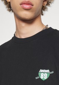 Camiseta negra de algodón con cuello redondo y un logo bordado en verde y blanco en el lado izquierdo, etiquetado como "WIP 89."