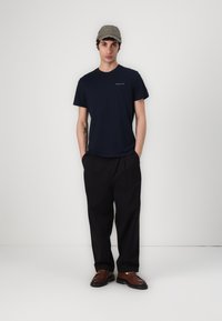 T-shirt en coton bleu marine associé à un pantalon noir décontracté et des chaussures marron. La casquette de baseball est grise avec des accents brodés.