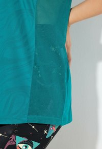 Camiseta atlética en color azul verdoso que presenta una tela suave y un panel lateral de malla. Combinada con leggins negros estampados que muestran gráficos coloridos.