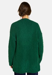 Frau mit braunem Haar in einem gedrehten Dutt, die goldene Kettenohrringe und einen strukturierten grünen Pullover mit langen Ärmeln trägt, von hinten gezeigt.