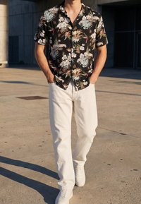 Homme marchant à l'extérieur portant une chemise noire à fleurs à manches courtes, un pantalon blanc et des baskets blanches, les mains dans les poches.
