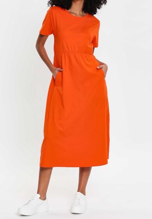 Frau trägt ein leuchtend orangefarbenes Midikleid mit kurzen Ärmeln und Taschen, kombiniert mit weißen Sneakers, steht vor einem schlichten Hintergrund.