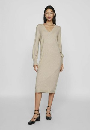 Robe en tricot beige avec un décolleté en V, des manches ballons longues et une coupe mi-longue. Portée avec des chaussures à talons noires dotées de brides de cheville.