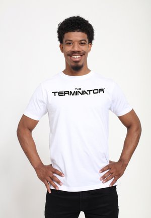 Hvid bomulds t-shirt med "THE TERMINATOR" trykt i fede sorte bogstaver. Kortærmet og med rund hals, båret med sorte bukser.