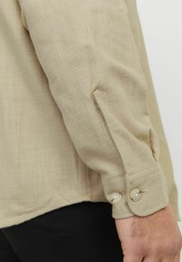 Beige langslievend shirt met een gestructureerde stof, met knoopmanchetten en een gebogen zoom, met gedeeltelijk zichtbare contrasterende donkere broek.