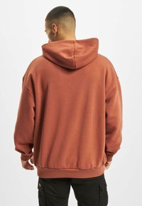 Sweat à capuche de couleur rouille, fabriqué en tissu doux, avec une coupe décontractée, des poignets et une taille côtelés. Texture lisse sans motifs visibles.