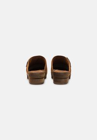 Sabots en daim marron avec une plateforme en bois, ouvert à l'arrière et à bout rond. Présentent un design cousu et une étiquette en cuir estampillée sur le côté.