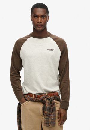 Long-sleeve shirt with a light gray body and brown raglan sleeves, featuring "SuperDry" logo. Paired with khaki pants and a plaid shirt tied around the waist.