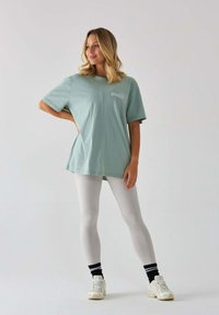 Jasnozielony oversize'owy T-shirt z krótkim rękawem i białym logo, połączony z białymi legginsami i jasnymi sneakersami. Gładkie tło.