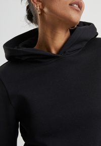 Sweat à capuche noir avec une grande capuche douce. Texture lisse et design ajusté, présentant un subtil éclat. De grandes boucles d'oreilles en or complètent le look.