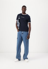  camiseta navy con ribete blanco que presenta el logotipo "GUESS JEANS", combinada con unos vaqueros azules de corte holgado y un bolso cruzado estampado. Zapatillas blancas.