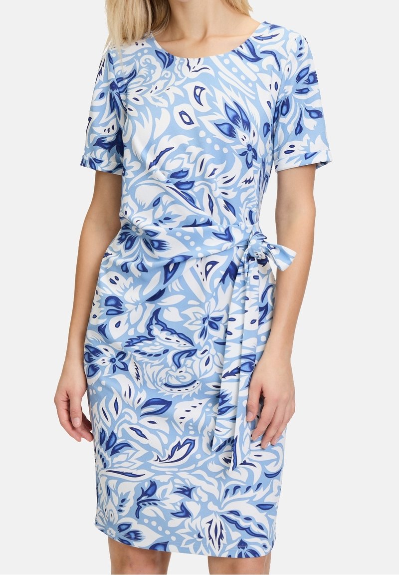 Femme portant une robe mi-longue à manches courtes avec un motif floral bleu et blanc et une ceinture en tissu nouée à la taille.