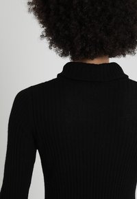 Pull noir côtelé à col roulé avec une texture douce, une coupe ajustée et un col montant. Le dos présente des détails subtils le long des coutures.