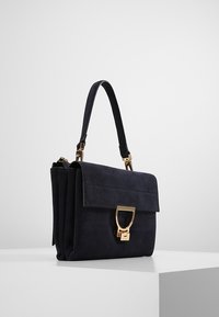 Czarna zamszowa torebka na ramię z złotym zapięciem i uchwytami, spoczywająca na białym pedestale na tle jednolitego jasnego tła.