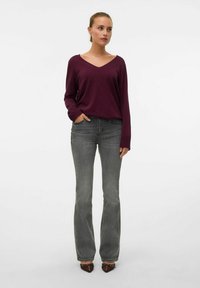 Top bordeaux a manica lunga con scollo a V abbinato a jeans grigi a vita alta a campana, con una texture liscia e dettagli di cucitura sottili. Scarpe marroni a punta.