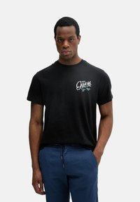 Camiseta de algodón negra con mangas cortas, cuello redondo y estampado gráfico en blanco de "Guess" en el pecho. Combinada con pantalones azules.