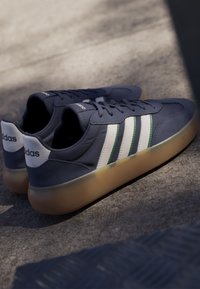 Par de zapatillas Adidas azul oscuro con rayas blancas y suelas color goma colocadas sobre una superficie gris texturizada a la luz del sol.