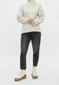 Pull en maille beige clair avec un col haut et un motif texturé. Associé à un jean gris foncé ajusté et des bottes à lacets couleur crème.
