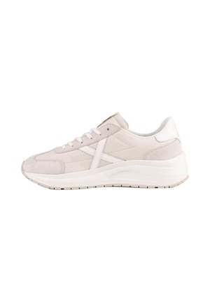 Zapatilla deportiva de color beige claro con cordones blancos, suela blanca y una franja blanca en forma de X en el lateral, mostrada de perfil sobre fondo blanco.