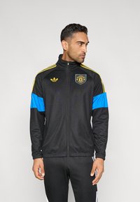 Melna sporta jaka ar dzeltenām svītrām, ziliem akcentiem, rāvējslēdzēju priekšpusē un izšūtu logotipu. Izgatavota no gludas auduma ar brīvu piegriezumu.