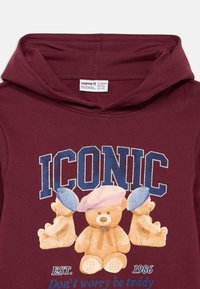 Sweatshirt bordeaux com o texto "ICÔNICO", três ursos de peluche a usar boinas azuis e rosas, e o lema "Não te preocupes, sê urso".