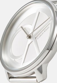 Srebrna ura iz nerjavečega jekla s krožno številčnico, ki ima brušeno belo ploščico in minimalističen dizajn, označena s "Calvin Klein."